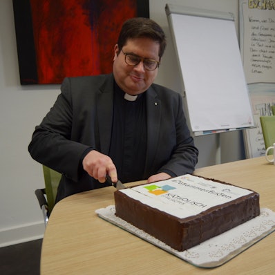 1Pfarrer Tobias Hopmann schneidet den Kuchen  an, der das Logo der neuen pastoralen Einheit Euskirchen zeigt.
