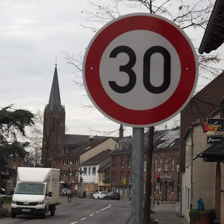 Ein Tempo-30 Schild steht an einer Straße, im Hintergrund sind Häuser und eine Kirche zu sehen.