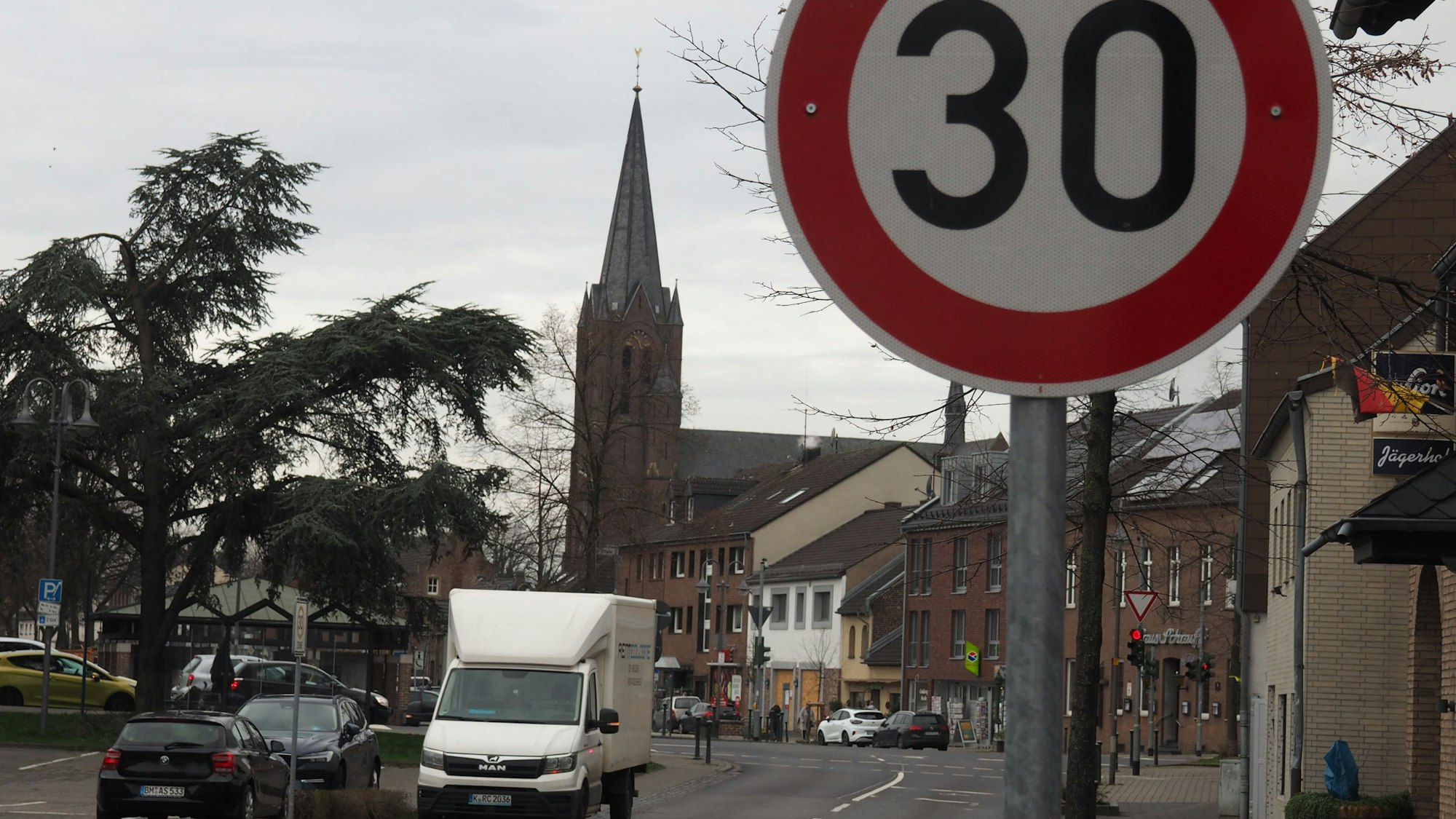 Ein Tempo-30 Schild steht an einer Straße, im Hintergrund sind Häuser und eine Kirche zu sehen.