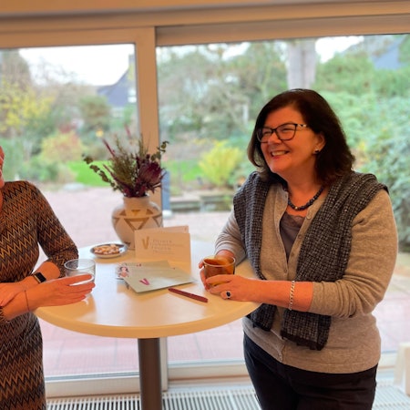 Eine blonde und eine dunkelhaarige Frau mittleren Alters stehen links und rechts vor einer Fensterfront, durch die man in einen Garten sieht, an einem Stehtisch.