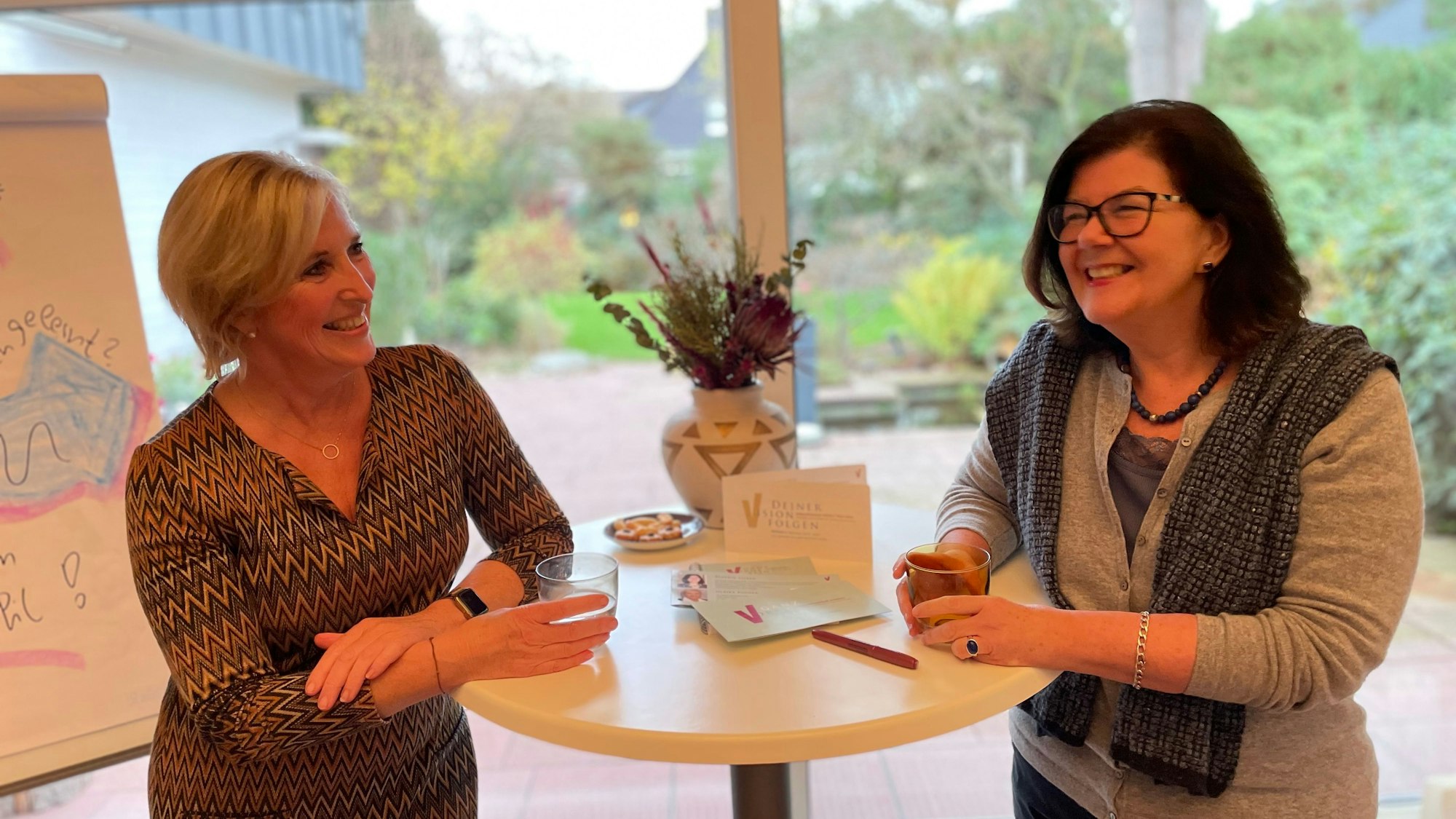 Eine blonde und eine dunkelhaarige Frau mittleren Alters stehen links und rechts vor einer Fensterfront, durch die man in einen Garten sieht, an einem Stehtisch.