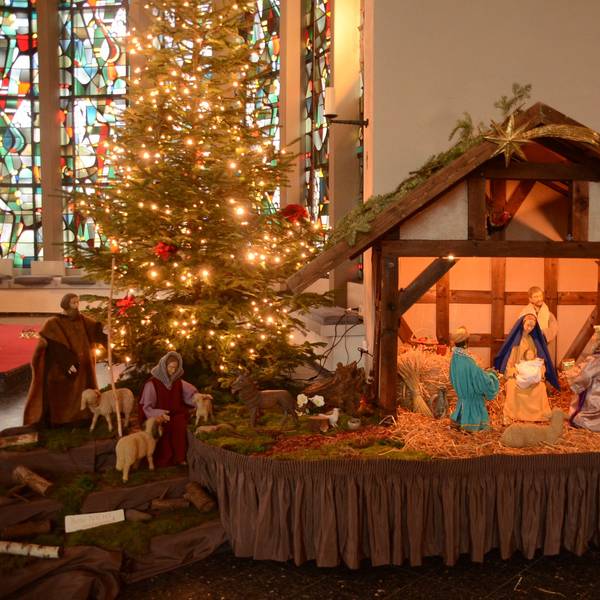 Krippe mit erleuchtetem Weihnachtsbaum vor Buntglasfenter links im Bild.