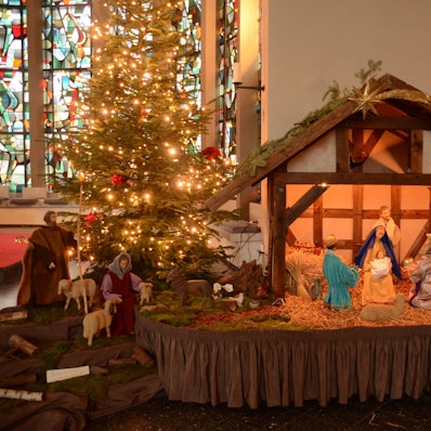 Krippe mit erleuchtetem Weihnachtsbaum vor Buntglasfenter links im Bild.