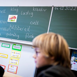 Drei Kinder in Schwarz sitzen in einem Klassenraum vor einer Tafel, die mit Adjektiven beschrieben ist.