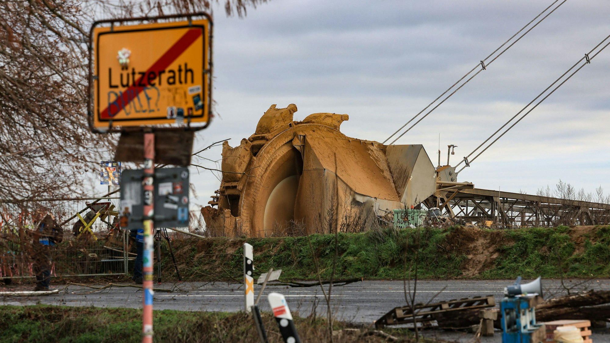 Im Bildvordergrund ist das Ortsschild von Lützerath zu sehen; dahinter, durch eine zweispurige Straße getrennt, der Arm eines riesigen Schaufelradbaggers, der, am Rande des Braunkohletagebaus Garzweiler, Erdreich schaufelt.