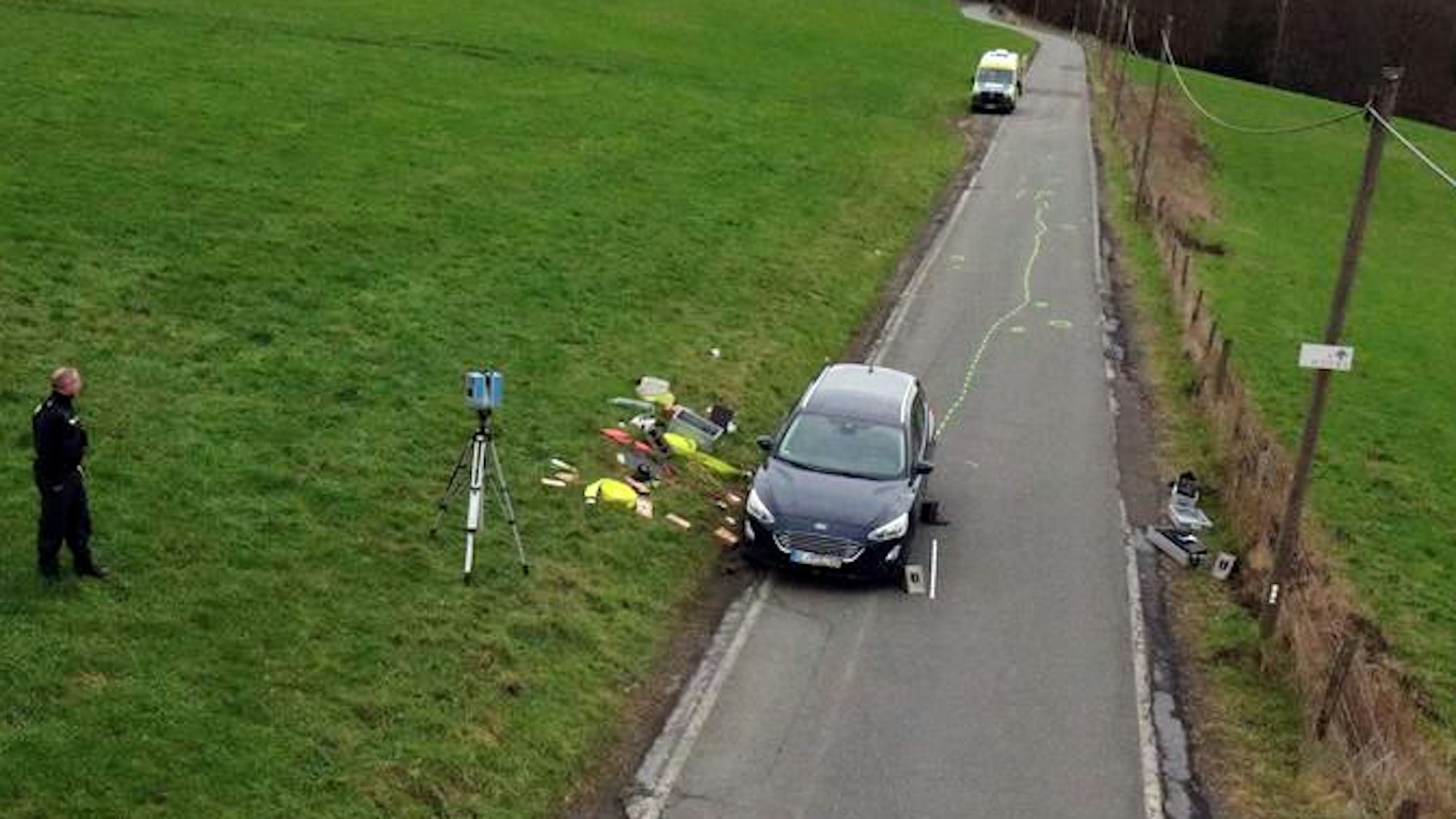 Ein Autor steht auf einer Straße, rund herum sind Unfallmarkierungen sowie ein BEamter des Unfallaufnahmeteams zu sehen.