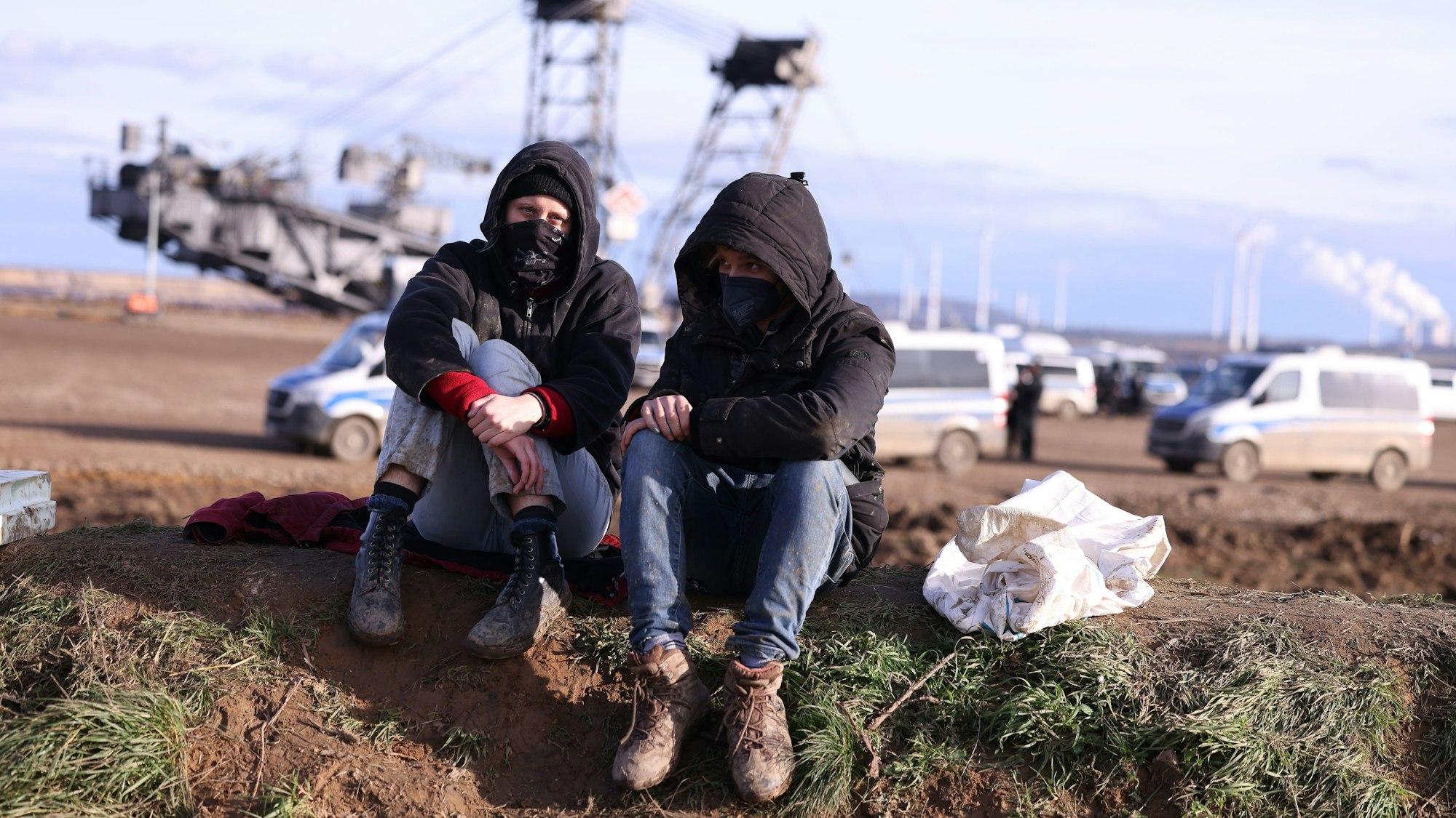 Zwei vermummte Klimaschutzaktivisten sitzen vor einem Schaufelradbagger am Rand des Braunkohledorfes Lützerath auf dem Boden. Im Hintergrund stehen mehrere Polizeifahrzeuge.