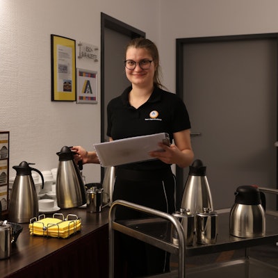 Eine schwarz gekleidete junge Frau stellt Kaffeekannen ab. Sie hält ein Klemmbrett in der Hand.