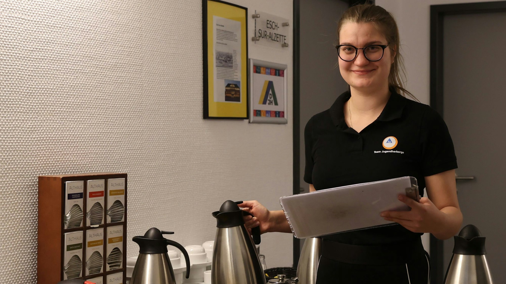 Eine schwarz gekleidete junge Frau stellt Kaffeekannen ab. Sie hält ein Klemmbrett in der Hand.