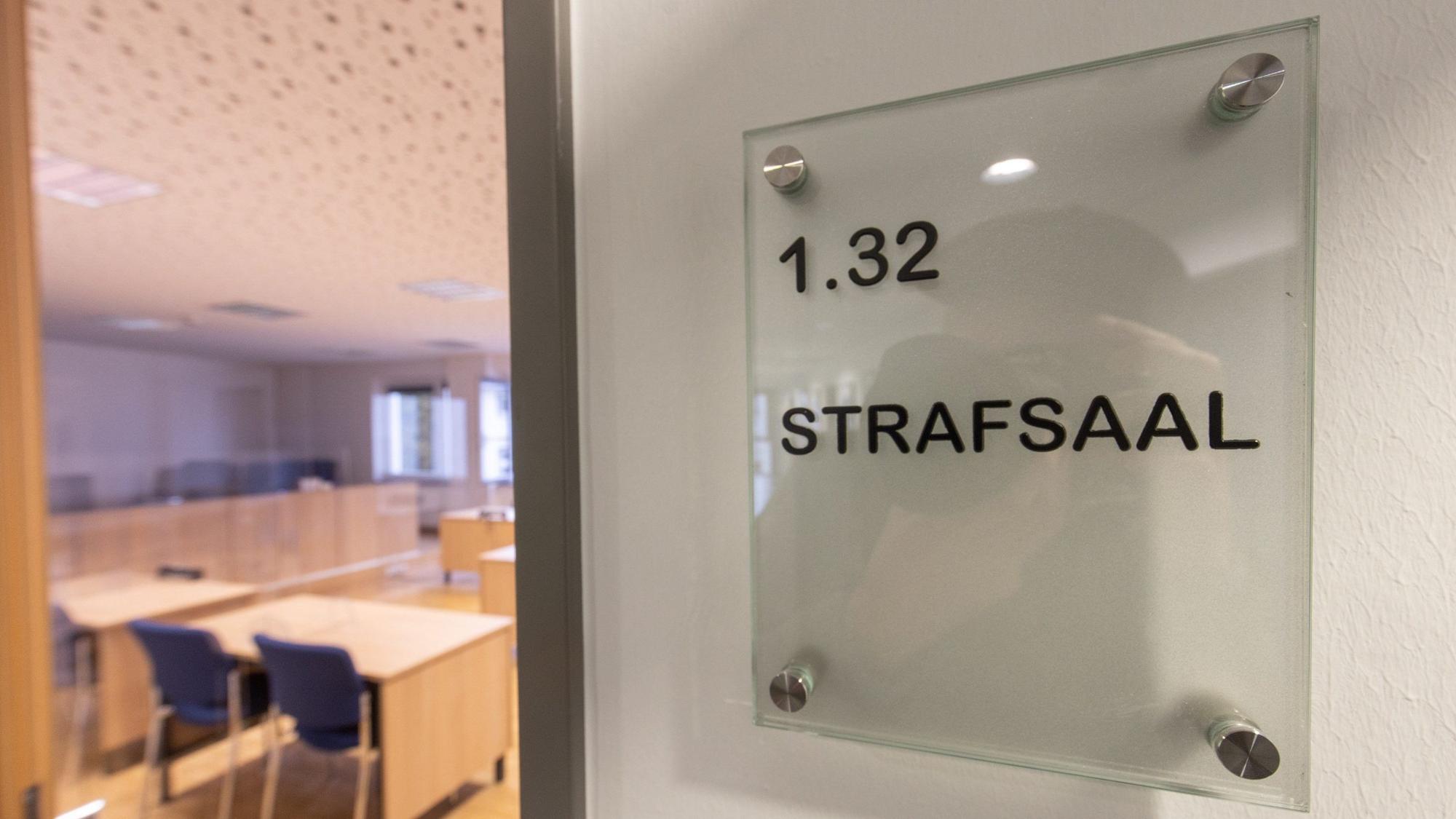 Die Tür eines Gerichtssaals steht einen Spalt offen. Im inneren sind leere Stühle und Tische zu sehen.