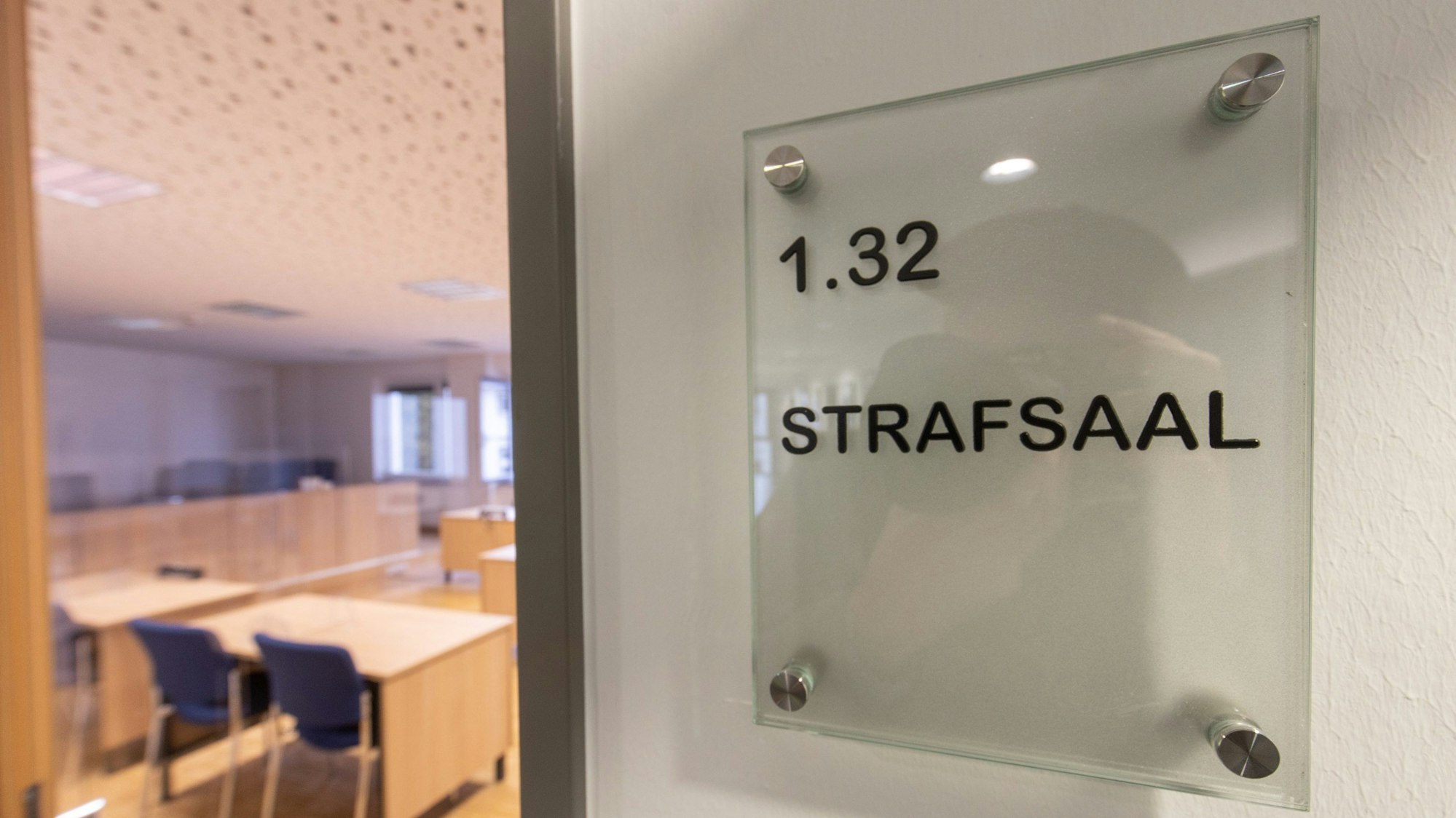 Die Tür eines Gerichtssaals steht einen Spalt offen. Im inneren sind leere Stühle und Tische zu sehen.