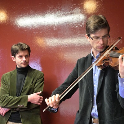 Roman Salyutov steht an einer Wand hinter Alexander Lifland, der auf der historischen Geige spielt.