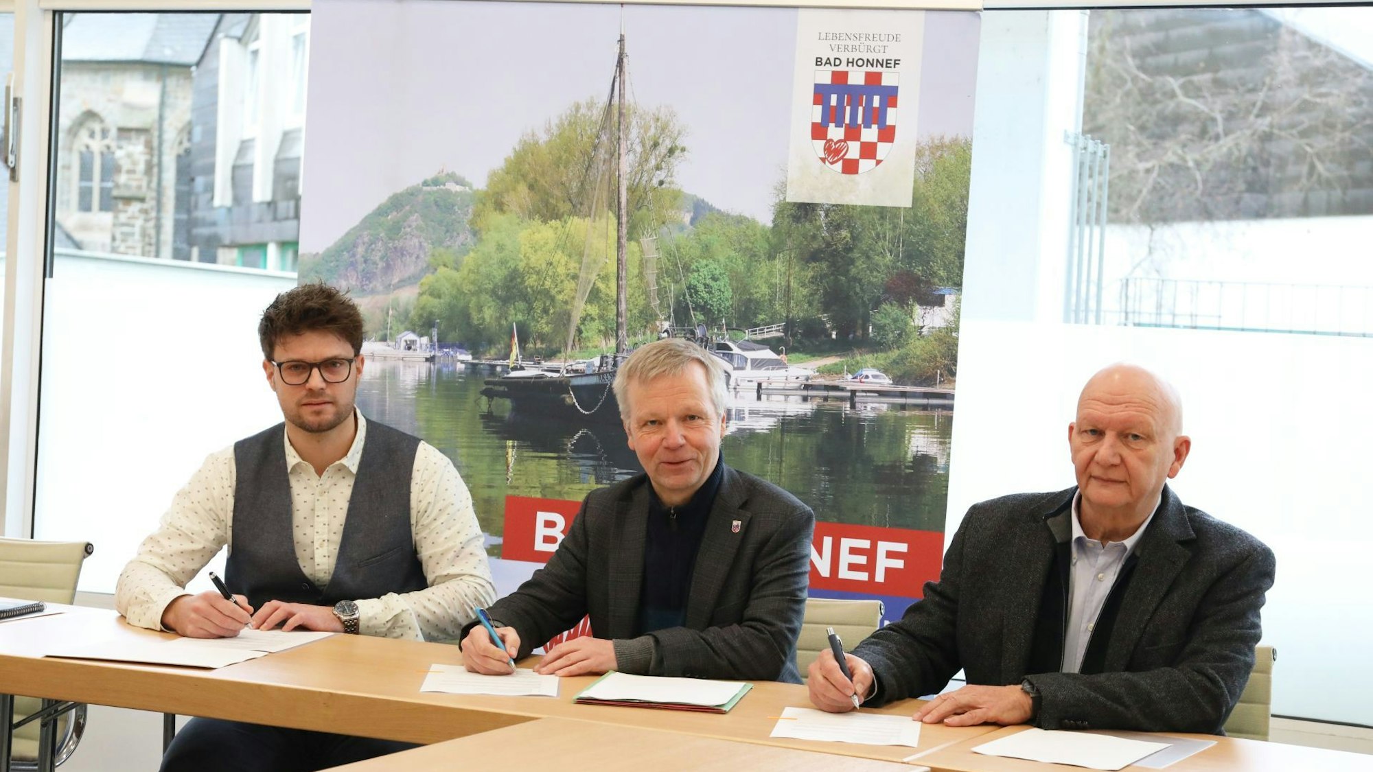 Drei Männer sitzen an einem Tisch, halten Kugelschreiber in den Händen und haben die Vereinbarung vor sich liegen. Ein Bild im Hintergrund zeigt den Aalschokker Aranka und den Drachenfels.