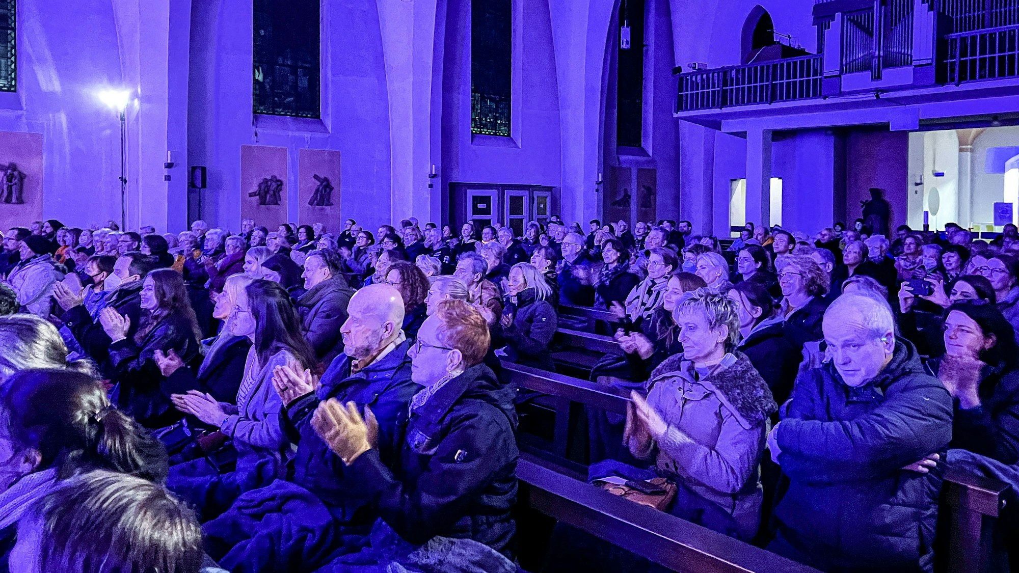 Auf den blau angestrahlten Kirchenbänken sitzen mehrere hundert Menschen und geben Applaus.