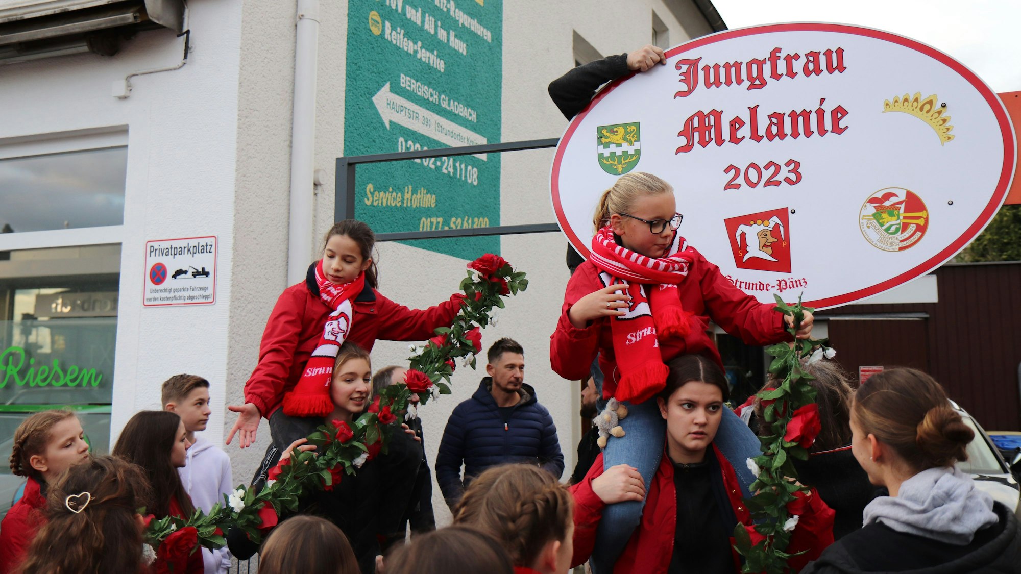 Zwei junge Mädchen sitzen auf Schultern von zwei jungen Frauen und schmücken mit Blumenketten das Haus der designierten Jungfrau.