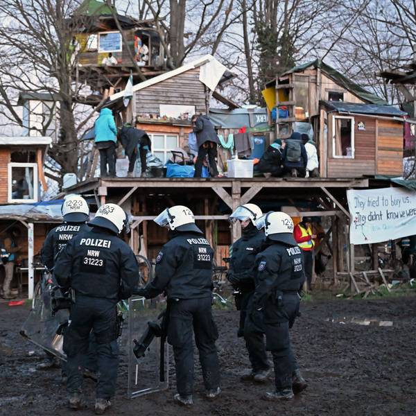 Polizisten mit Ausrüstung und Helmen stehen in einer Gruppe vor Bretterhütten, die Aktivisten aufgebaut haben. Auf einem Podest an den Hütten befinden sich mehrere Aktivisten.