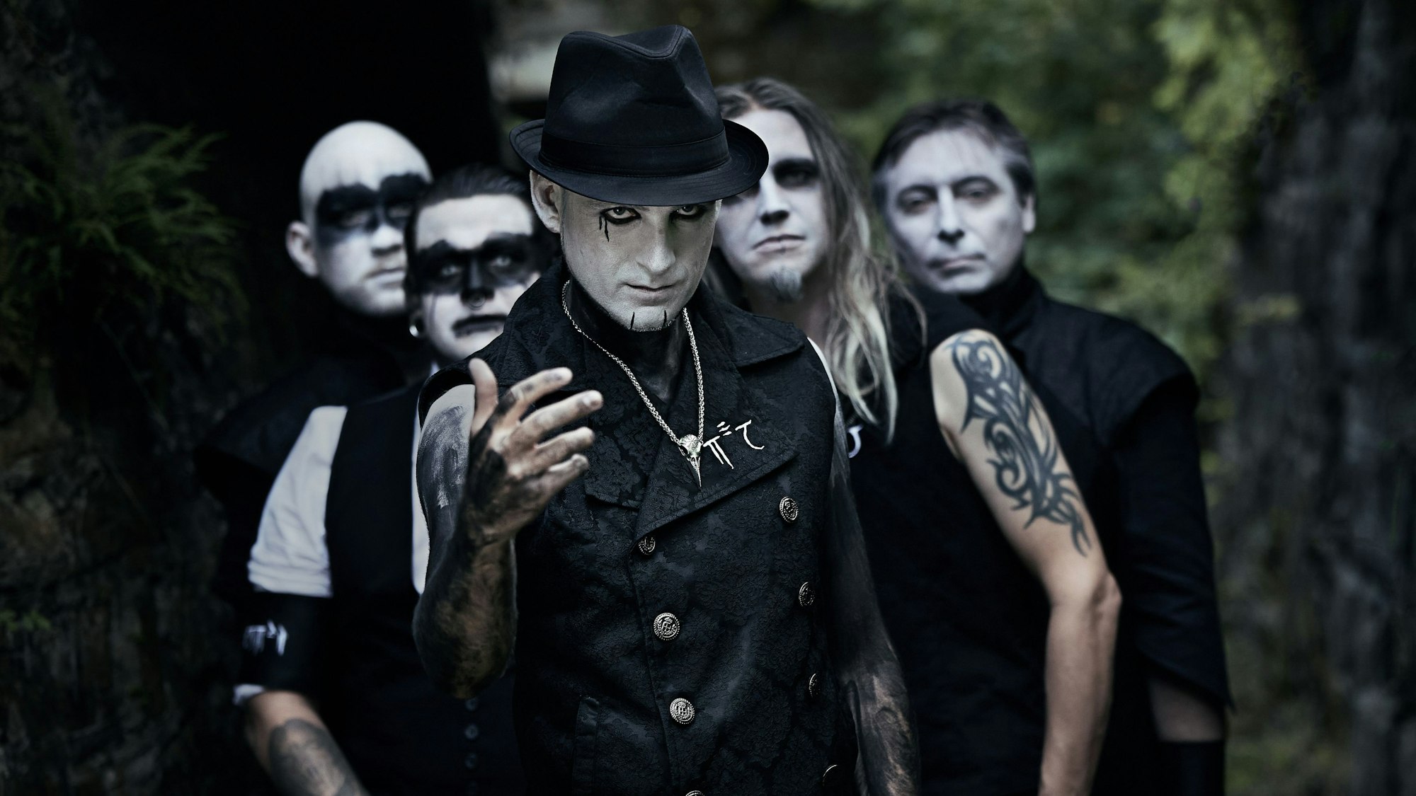 Fünf schwarz gekleidete Männer haben sich die Gesichter weiß geschminkt und mit schwarzer Farbe ihre Augen teilweise großflächig geschminkt. Einer der Männer ist am Oberarm tätowiert.