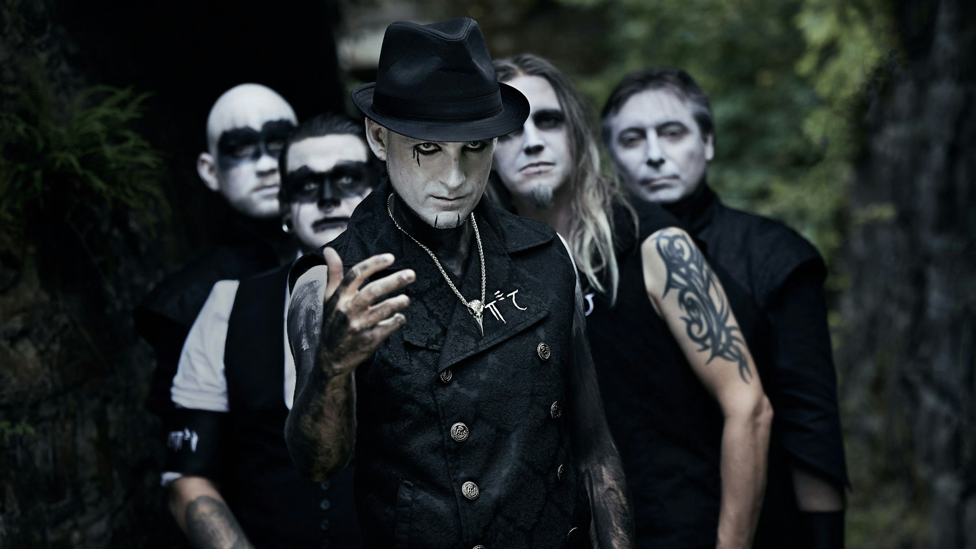 Fünf schwarz gekleidete Männer haben sich die Gesichter weiß geschminkt und mit schwarzer Farbe ihre Augen teilweise großflächig geschminkt. Einer der Männer ist am Oberarm tätowiert.