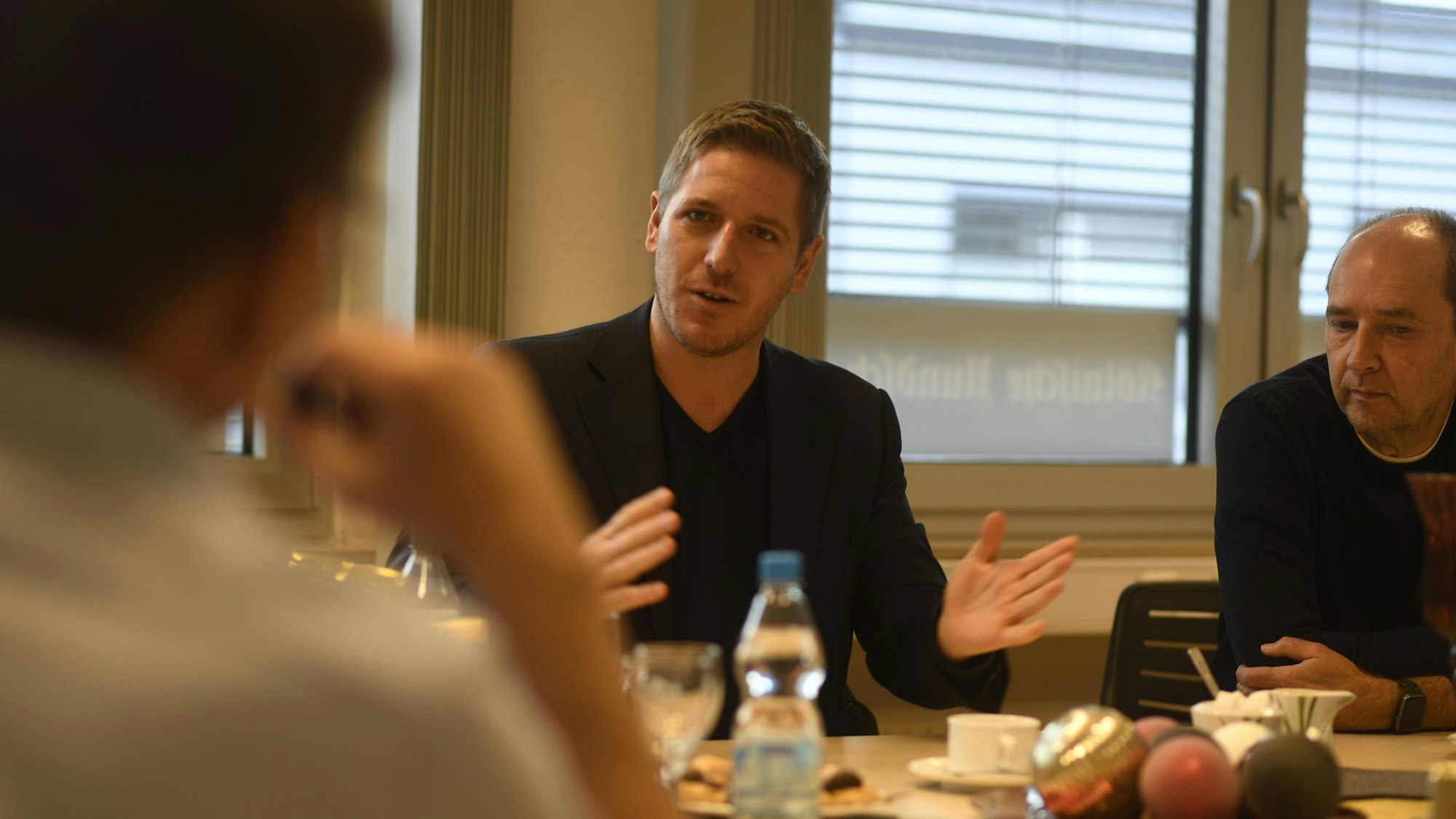 Drei Männer sitzen im Gespräch am Tisch, einer gestikuliert mit den Händen.