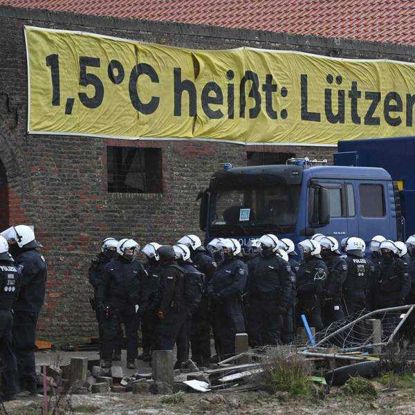 Ein Banner mit der Aufschrift "1,5°C heißt: Lützerath bleibt!" hängt an einem verlassenen Gebäude. Davor stehen Polizisten.