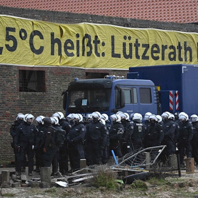 Ein Banner mit der Aufschrift "1,5°C heißt: Lützerath bleibt!" hängt an einem verlassenen Gebäude. Davor stehen Polizisten.