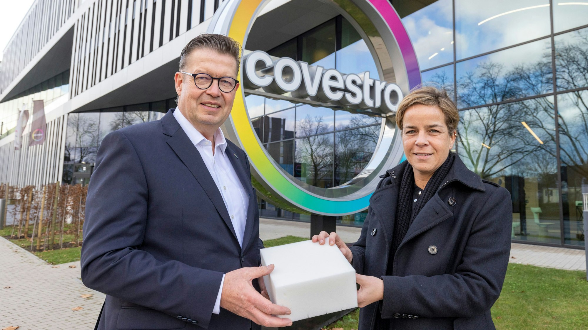 Produktionsvorstand Klaus Schäfer zeigt NRW-Wirtschaftsministerin Mona Neubaur vor der Covestro-Zentrale einen Würfel aus recyceltem Schaumstoff