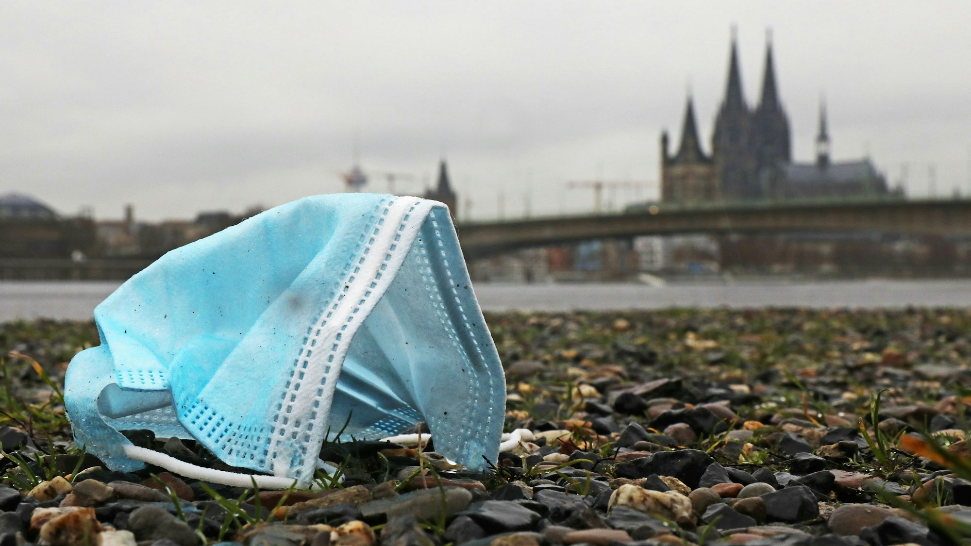 Eine weggeworfene Maske liegt am Rheinufer auf dem Boden. Im Hintergrund ist der Dom zu sehen. In Nordrhein-Westfalen galten strenge Regeln, um die Ausbreitung des Coronavirus zu verhindern.
