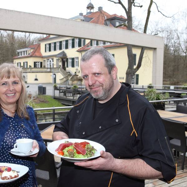 Marlene Jablonski-Reichelt und ihr Koch Mario Arke stehen neben Schloss Eulenbroich und halten Teller mit Essen darauf in den Händen.