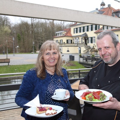 Marlene Jablonski-Reichelt und ihr Koch Mario Arke stehen neben Schloss Eulenbroich und halten Teller mit Essen darauf in den Händen.