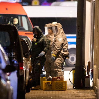Einsatzkräfte in Schutzmontur und mit Atemmasken stehen auf dem Bürgersteig in Castrop-Rauxel neben Einsatzwagen.