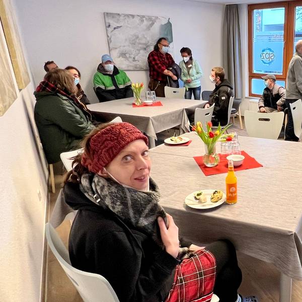 In einem Gemeinschaftsraum sitzen Menschen an drei größeren Tischen zusammen, einige tragen Masken.