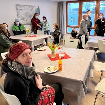 In einem Gemeinschaftsraum sitzen Menschen an drei größeren Tischen zusammen, einige tragen Masken.