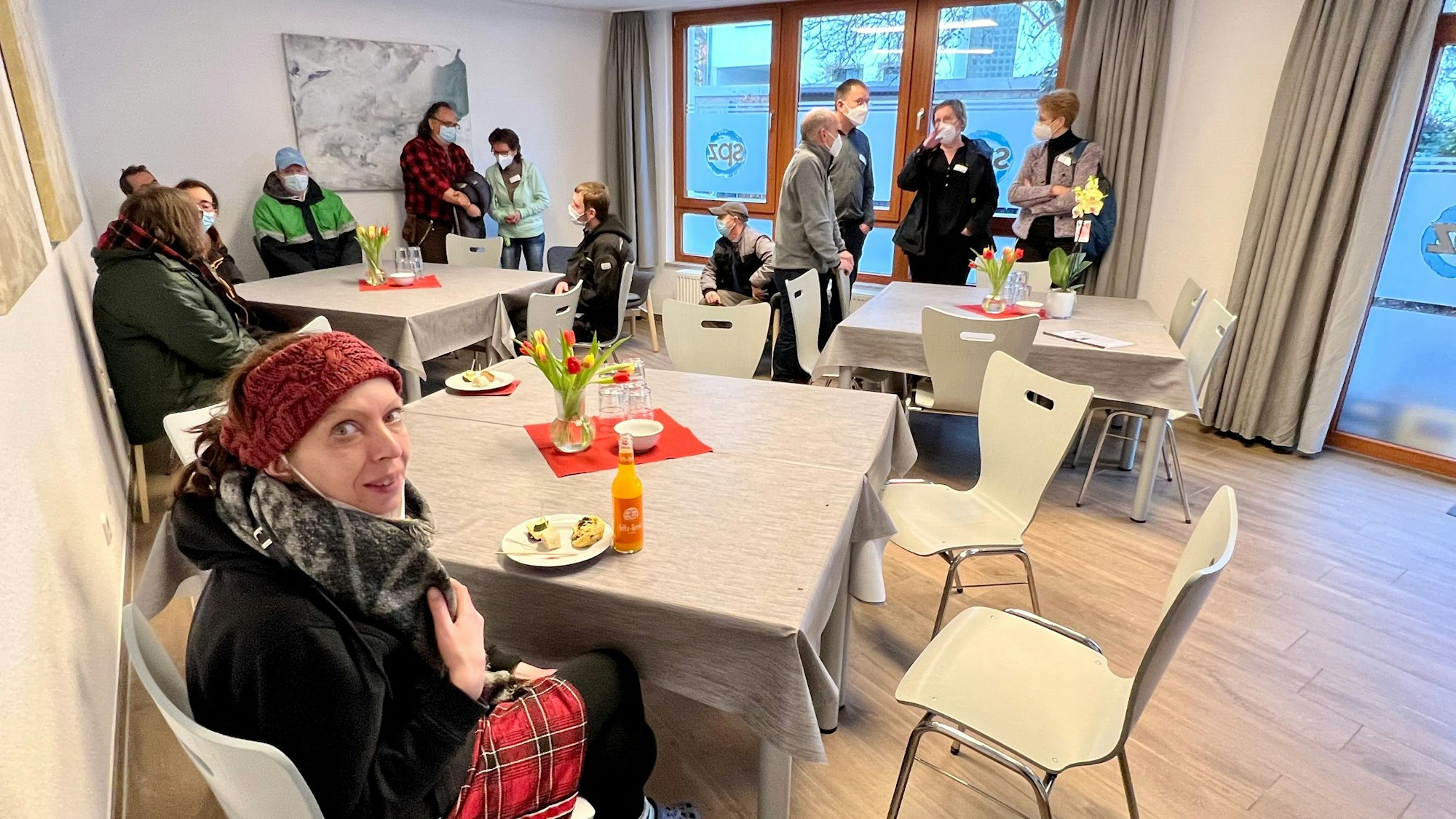 In einem Gemeinschaftsraum sitzen Menschen an drei größeren Tischen zusammen, einige tragen Masken.