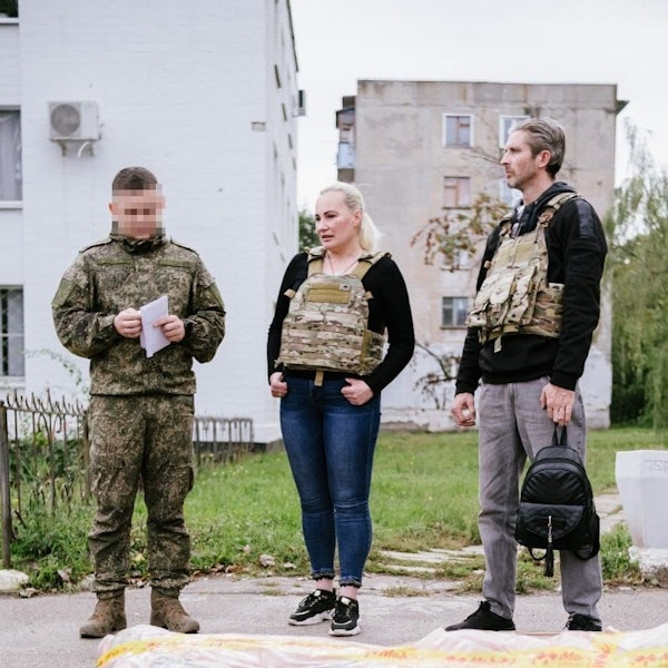 Elena Kolbasnikova und Max Schlund stehen mit schusssicherer Weste auf einem Gehweg zwischen Plattenbauten. Schlund hat einen schwarzen Frauenrucksack in der Hand. Links von ihnen steht ein Soldat.