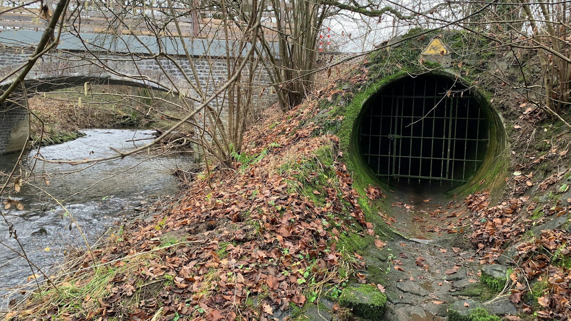 Neben einem Bach, über den eine Brücke führt, ist eine vergitterte Öffnung zu sehen.