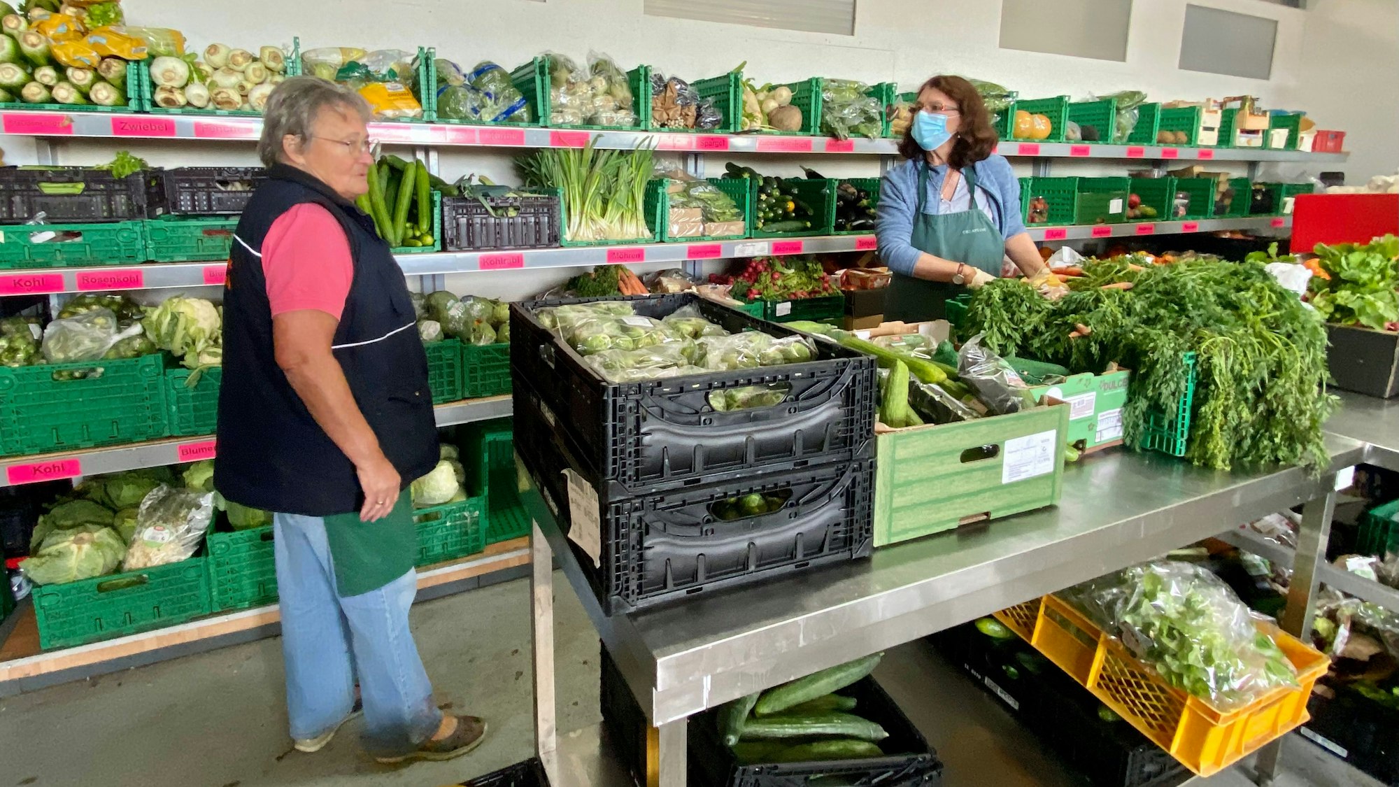 Margret Welsch und Helmi Dreifke sortieren Lebensmittel in der Euskirchener Tafel. Im Hintergrund sind prall gefüllte Regale mit Lebensmittelkisten zu sehen.