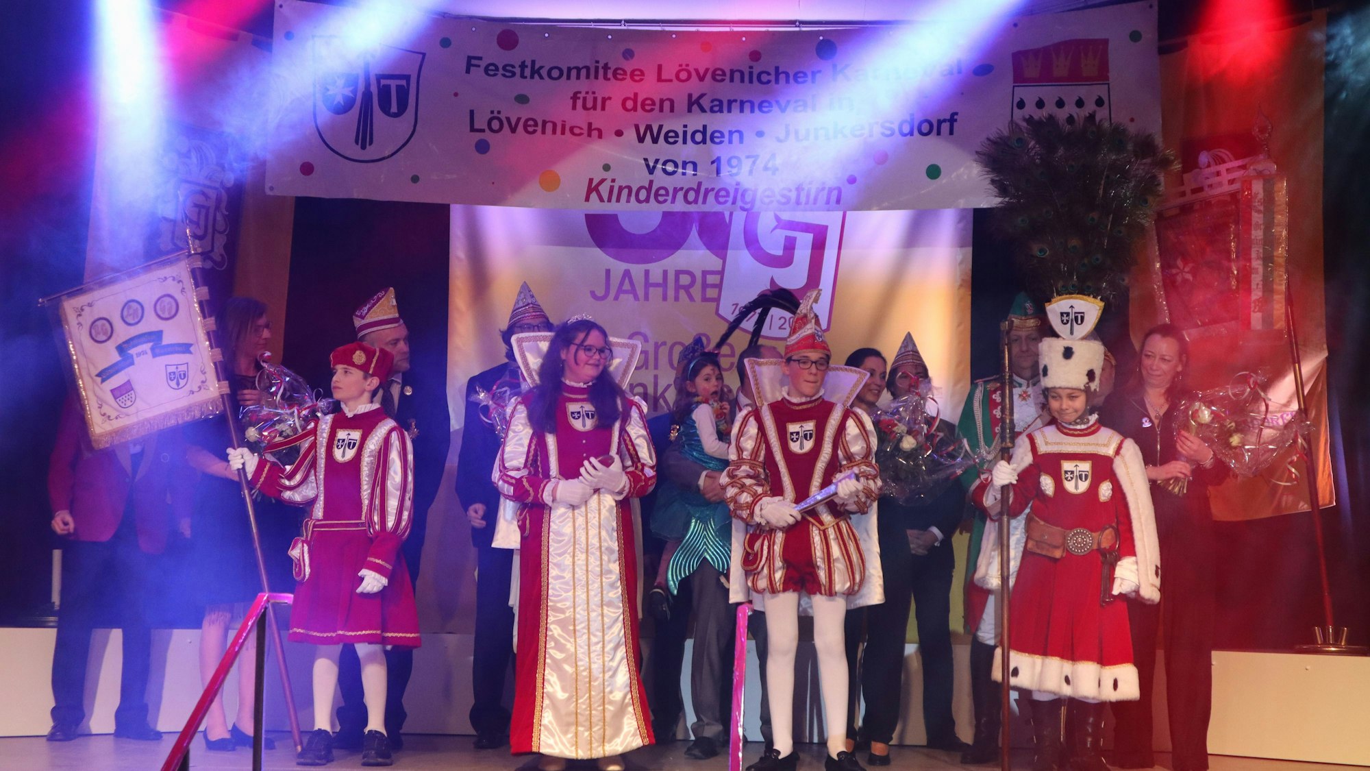Vier Kinder in rot-weißen Karnevalstrachten stehen auf der Bühne. Hinter ihnen sind noch einige Erwachsene im Kostüm zu sehen.