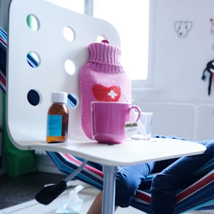 Fieberthermometer, Hustensaft, Wärmflasche und eine Tasse Tee stehen auf einem Stuhl, während ein Kind im Hintergrund in einer Hängematte liegt (Gestellte Szene).