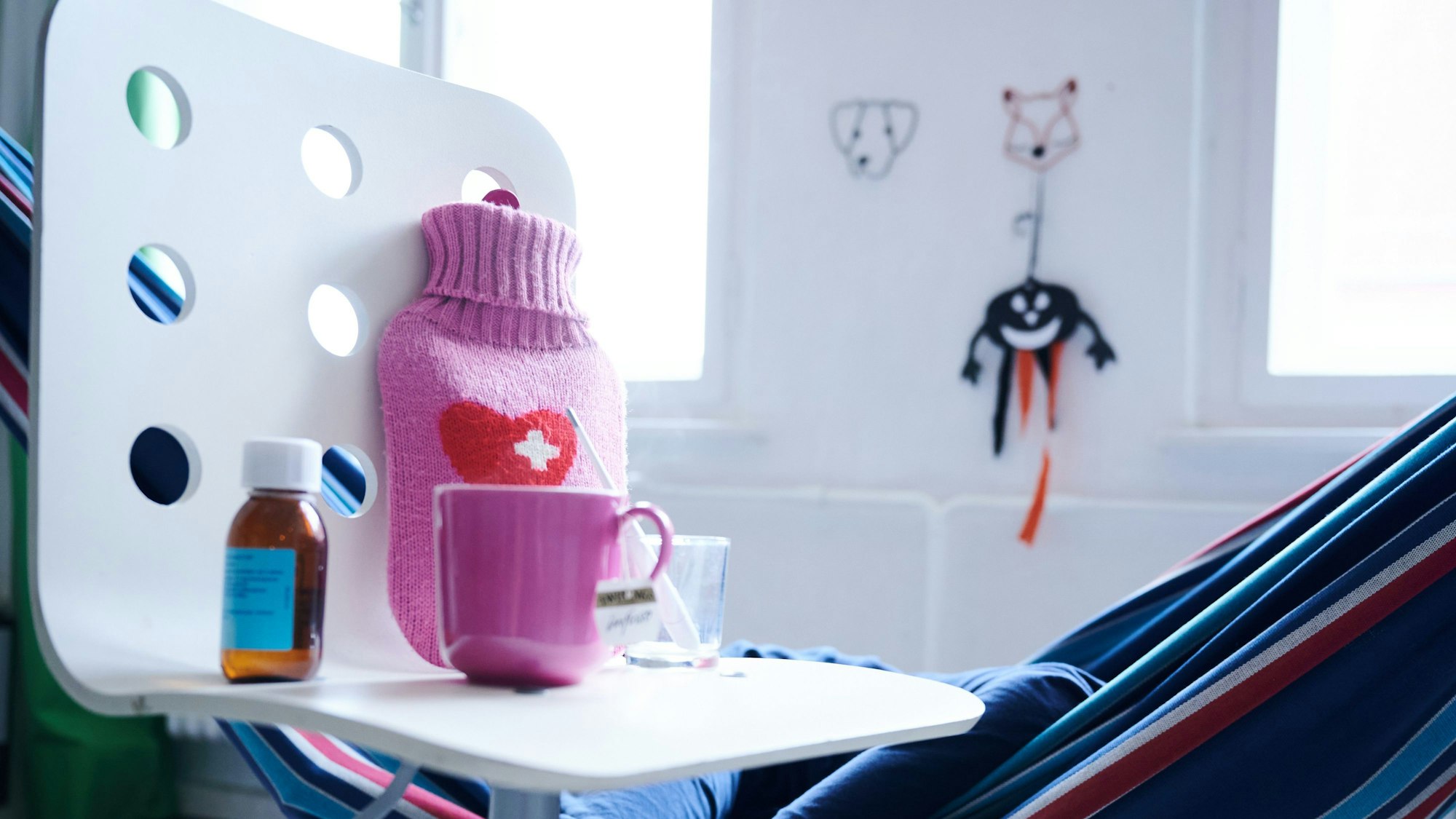 Fieberthermometer, Hustensaft, Wärmflasche und eine Tasse Tee stehen auf einem Stuhl.