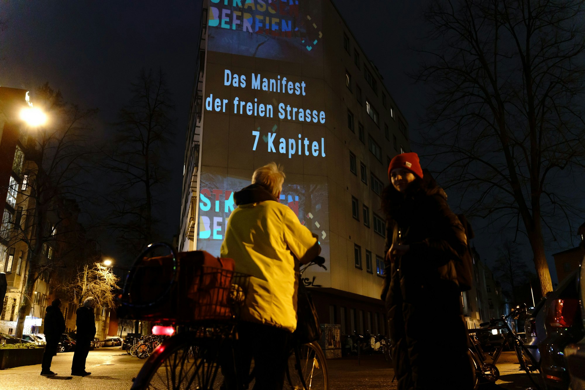 Organisatorin Theresa Pfaff des Projekts „Strassen befreien“ im Gespräch mit einer Fahrradfahrerin am Sudermanplatz in Köln. Hinter ihnen ist auf einer Hauswand in großen Buchstaben projiziert „Strassen befreien / Das Manifest der freien Straße / 7 Kapitel“.