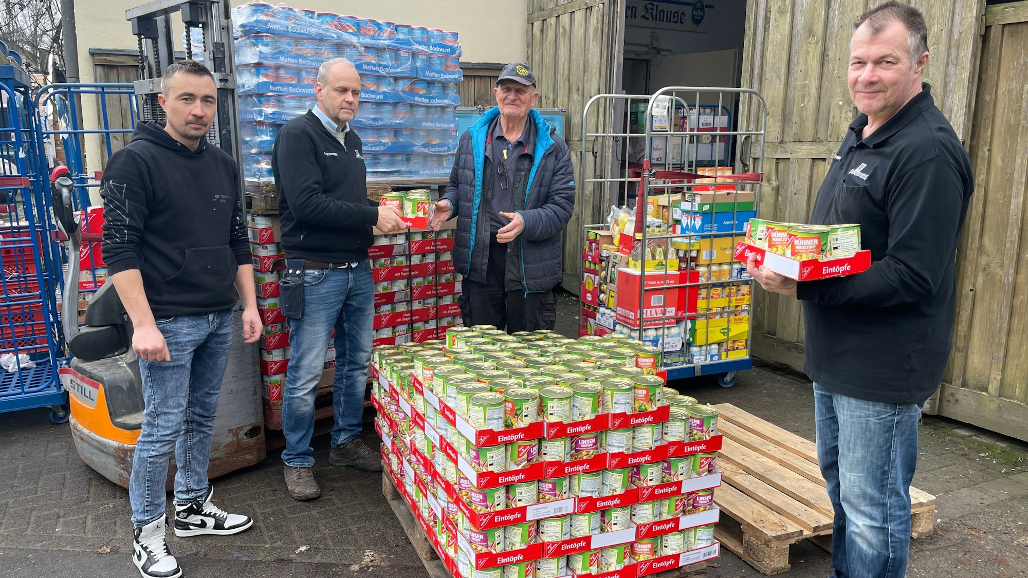 Vier Männer stehen vor Paletten voll mit Dosen voller Lebensmittel.