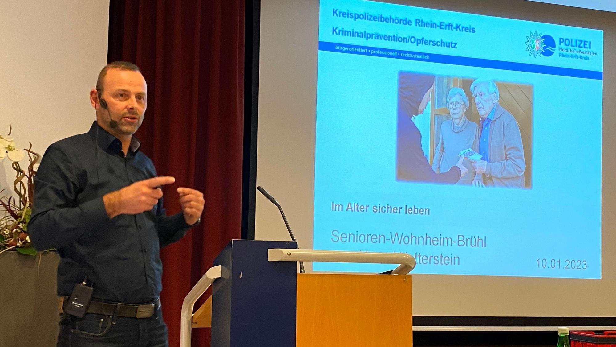 Kriminalhauptkommissar Guido Kümpel steht am Rednerpult. Hinter ihm ist eine PowerPoint-Präsentation an die Wand projiziert.