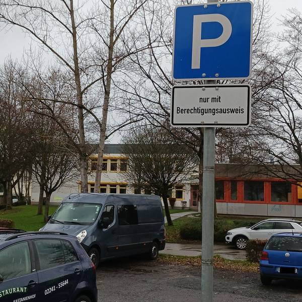 Einige Fahrzeuge parken auf einem asphaltierten Platz, hinter dem kahle Bäume und das graue Flachdachgebäude des Schulzentrums Neuenhof stehen.