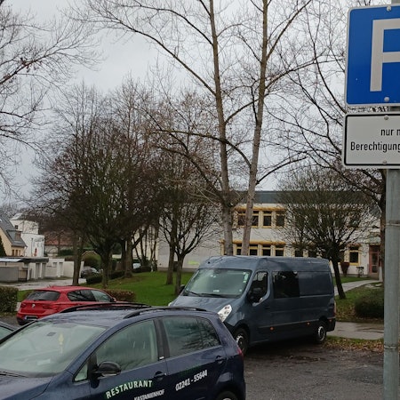 Einige Fahrzeuge parken auf einem asphaltierten Platz, hinter dem kahle Bäume und das graue Flachdachgebäude des Schulzentrums Neuenhof stehen.