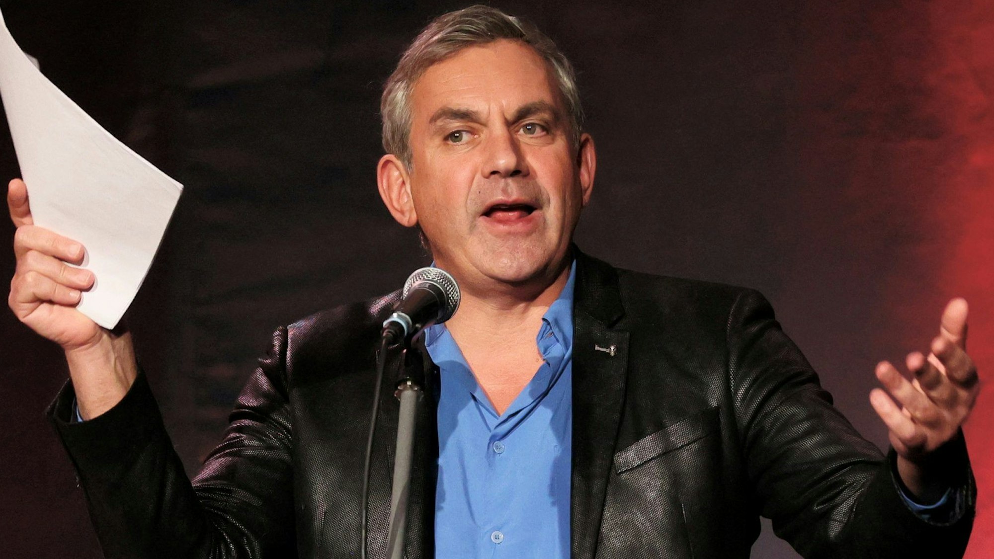 Wladimir Kaminer mit Zettel in der Hand an einem Tisch, auf dem ein Glas steht und ein Buch liegt.
