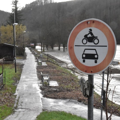 Ein Verbotsschild für Kraftfahrzeuge steht an einem schmalen asphaltierten Weg zwischen dem Fluss Sieg und einem Grünstreifen. Zwischen Sitzbänken sind die geschredderte Überreste von Vegetation zu erkennen.