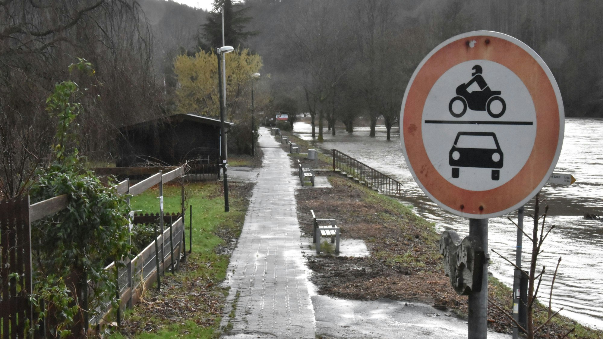 Ein Verbotsschild für Kraftfahrzeuge steht an einem schmalen asphaltierten Weg zwischen dem Fluss Sieg und einem Grünstreifen. Zwischen Sitzbänken sind die geschredderte Überreste von Vegetation zu erkennen.