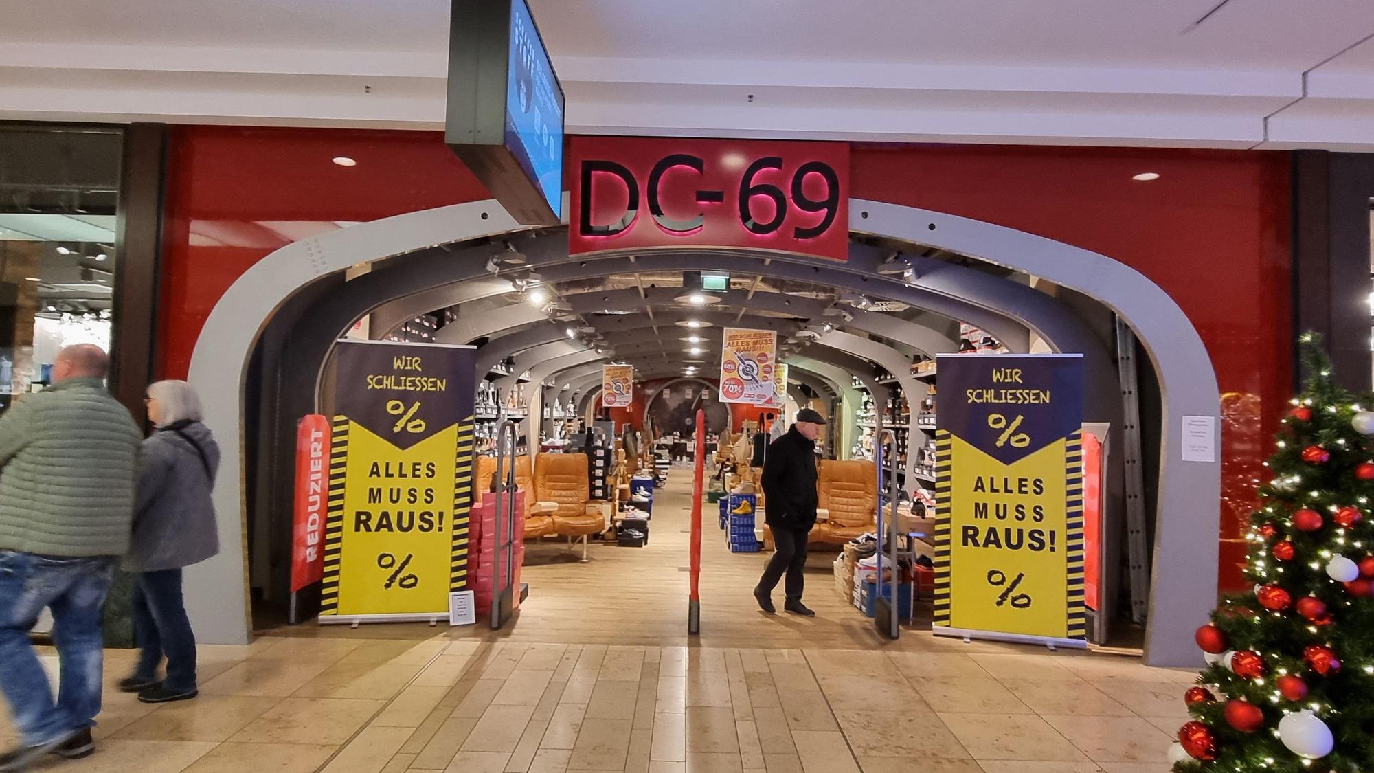 Der Eingang des Schuhgeschäfts DC-69. Links und rechts stehen Schilder mit „Wir schließen. Alles muss raus!“ Ein Mann schaut sich die Produkte an.