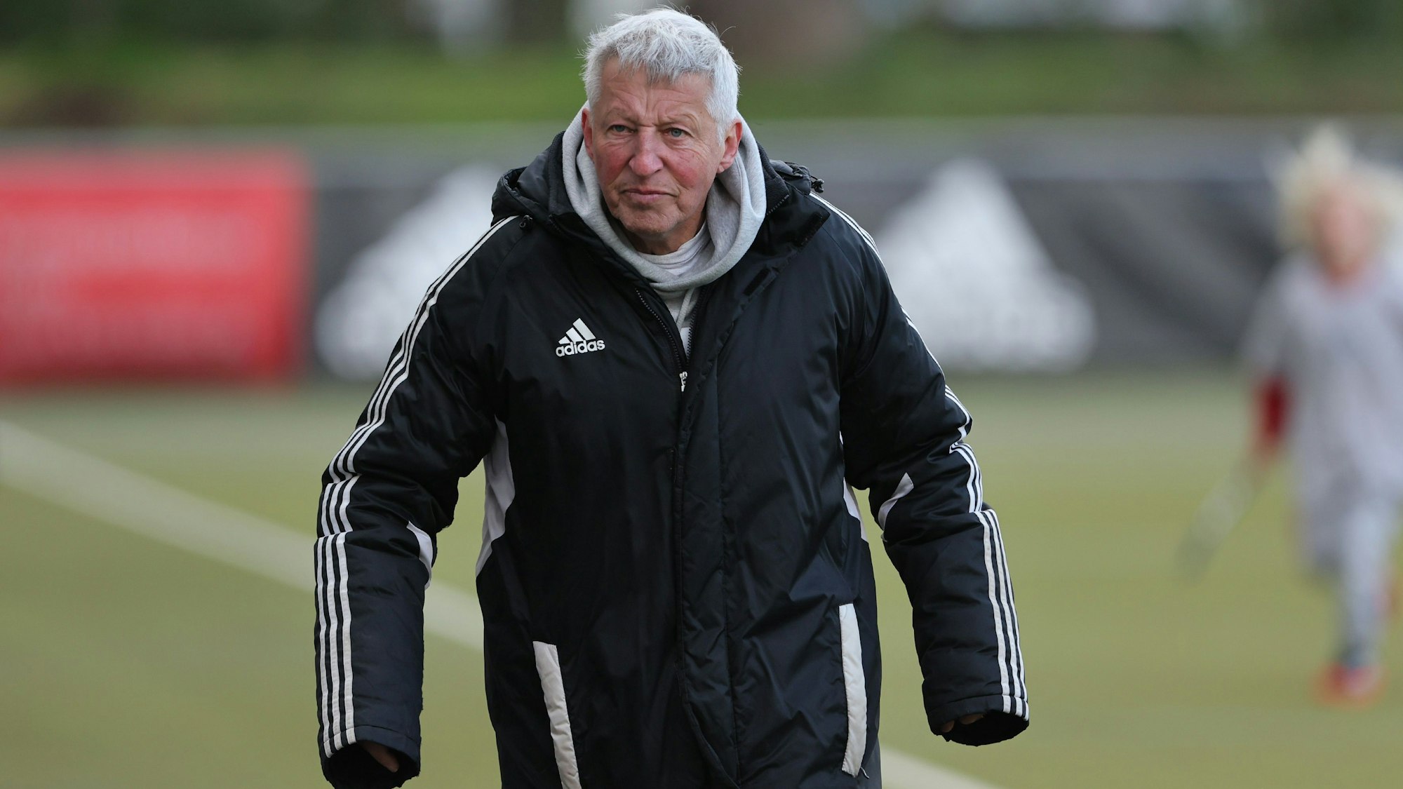 Ein Hockeytrainer steht am Spielfeldrand und hat die Hände in den Ärmeln seiner Jacke zum Aufwärmen versteckt.