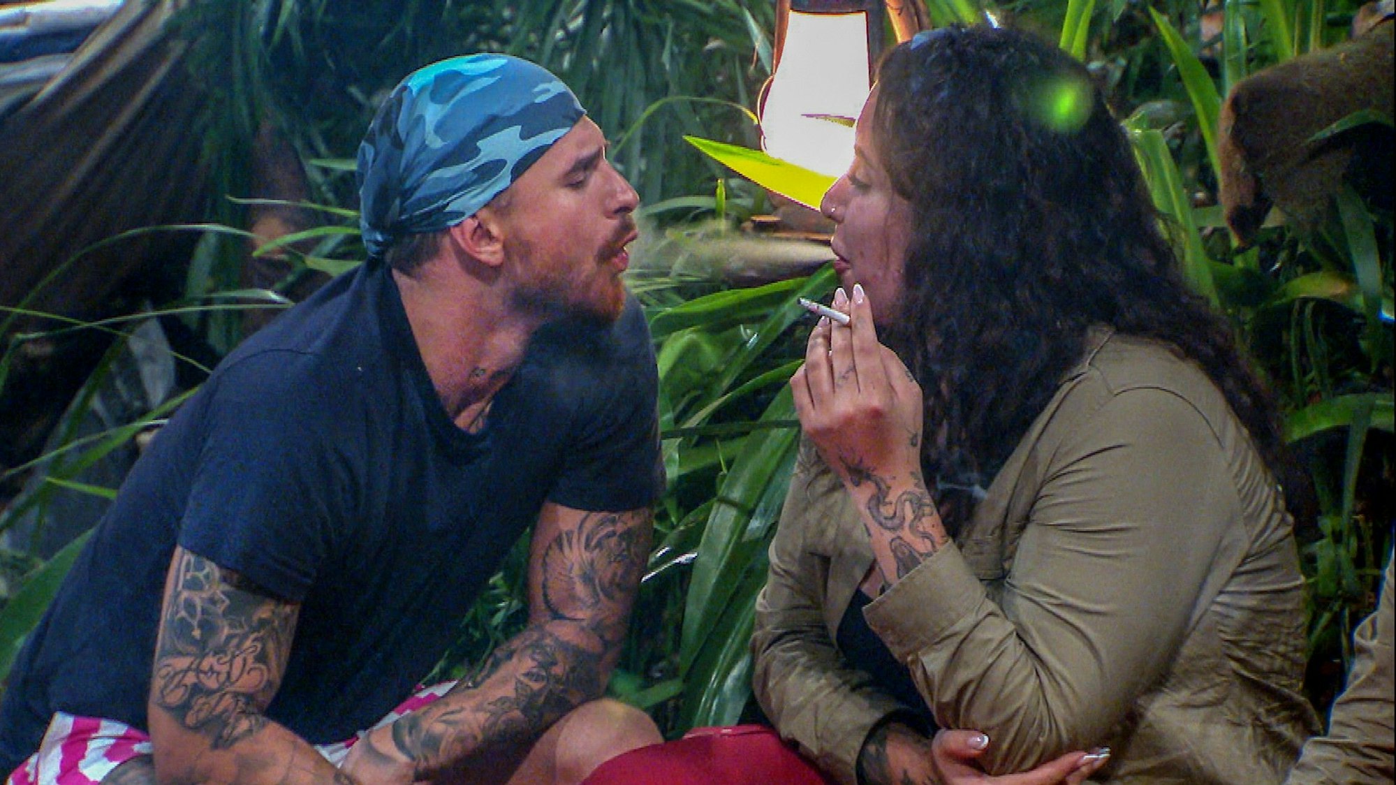 Dschungelcamp 2023: Gigi Birofio lässt sich von Jolina Mennen Rauch in den Mund blasen, während er an einem Stöckchen wie an einer imaginären Zigarette zieht.